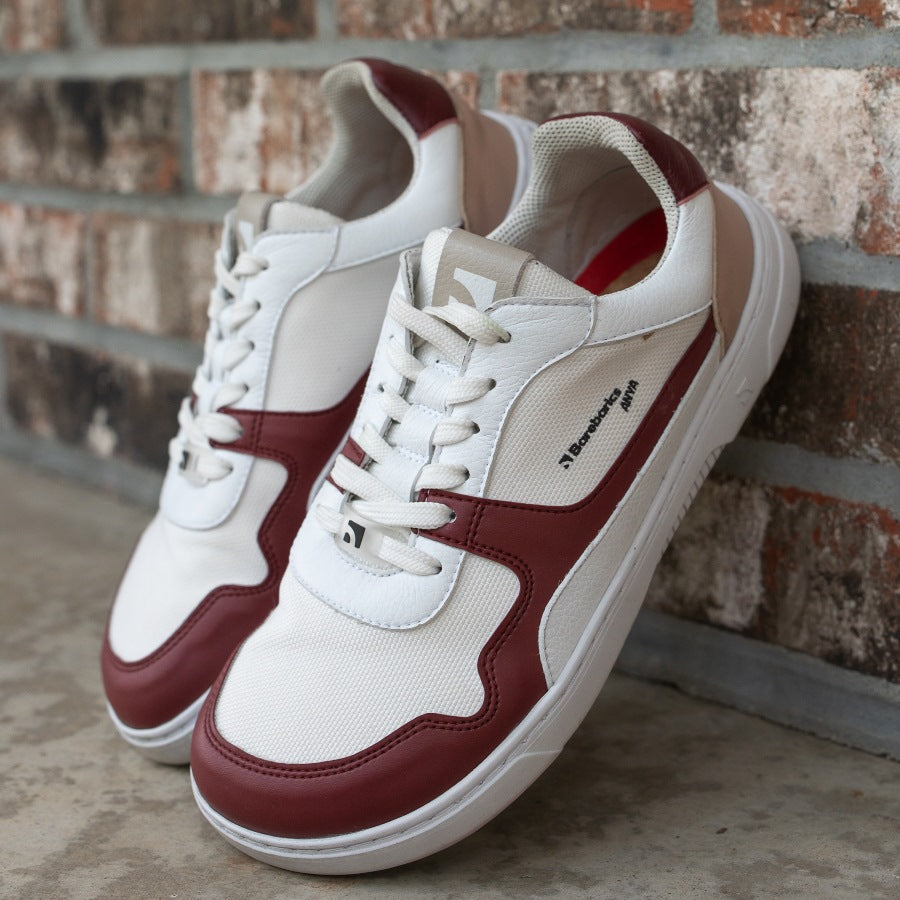 Barebarics Zing sneakers made with a leather upper and a rubber sole. The sneakers are white with burgundy accents around the front and side, and a tan accent over the heel. There are perforated spots on the top of the toe box and barebarics branding on the tongue and side. Both shoes are shown together from the front left, propped up on a brick wall. #color_burgundy-anya-exclusive