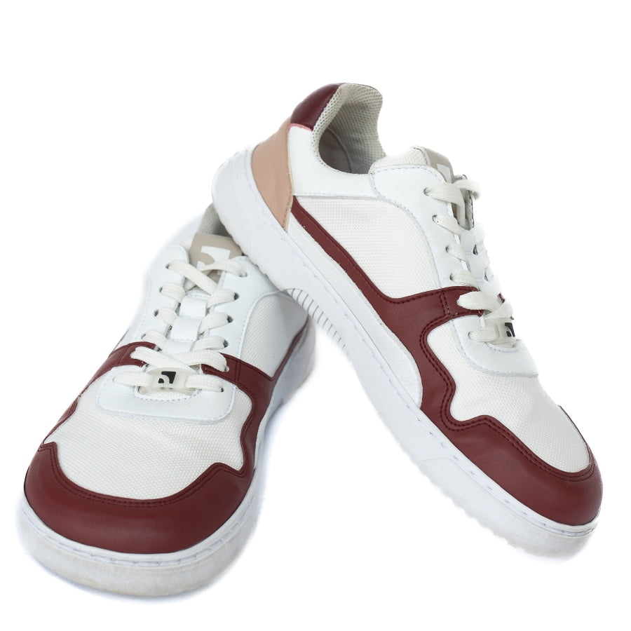 Barebarics Zing sneakers made with a leather upper and a rubber sole. The sneakers are white with burgundy accents around the front and side, and a tan accent over the heel. There are perforated spots on the top of the toe box and barebarics branding on the tongue and side. Both sneakers are shown from the front with the heel of the left shoe resting up on the tongue of the right shoe, on a white background. #color_burgundy-anya-exclusive