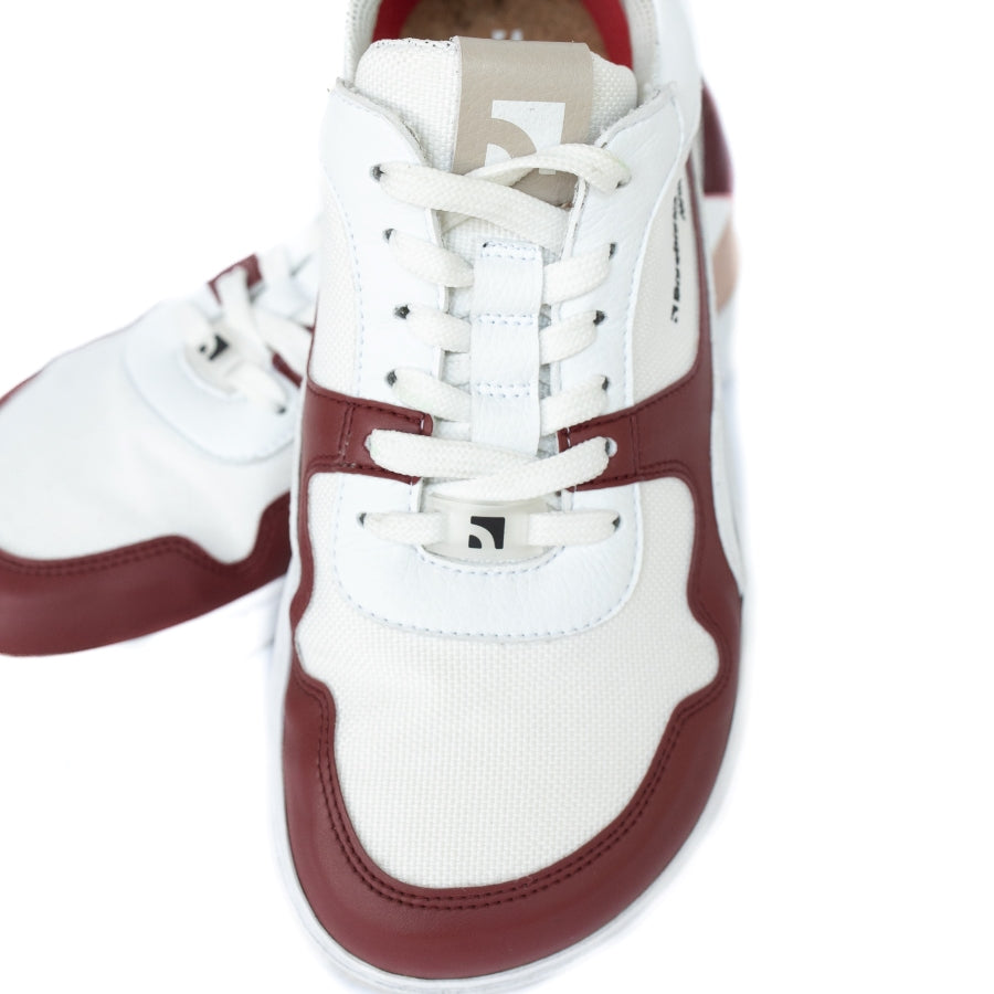 Barebarics Zing sneakers made with a leather upper and a rubber sole. The sneakers are white with burgundy accents around the front and side, and a tan accent over the heel. There are perforated spots on the top of the toe box and barebarics branding on the tongue and side. Both sneakers are shown from above with the left shoe resting atop the right shoe, on a white background. #color_burgundy-anya-exclusive