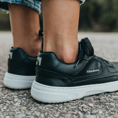 A photo of Barebarics Zing sneakers made with a leather upper and a rubber sole. The sneakers are a black color with perforated spots on the top of the toe box and barebarics branding on the tongue and side. Both sneakers are shown on a woman’s feet from the right with a view of her ankles down and a close up of the right heel. The woman is wearing cropped skinny blue jeans and is standing on asphalt. #color_black-white