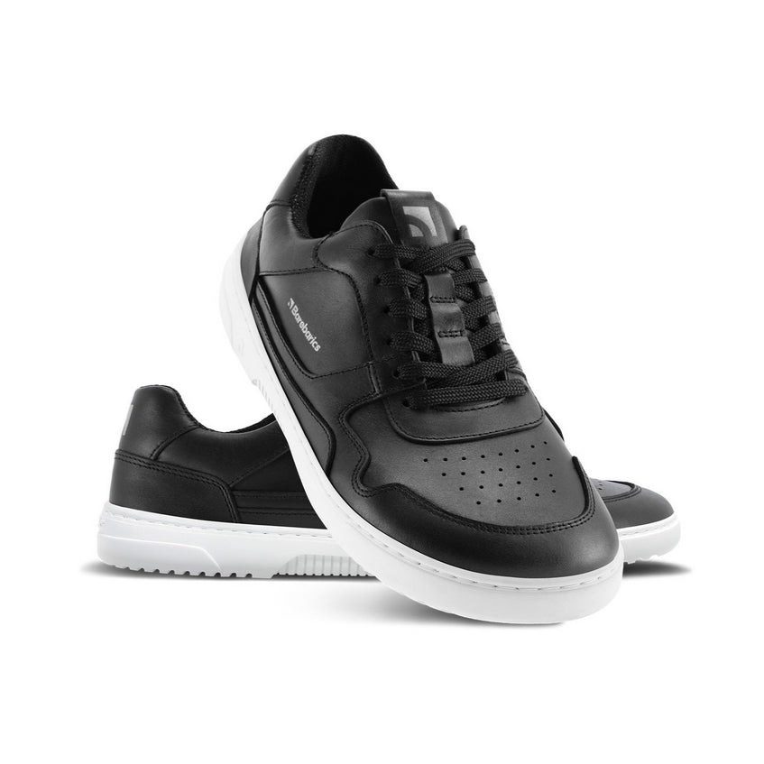 A photo of Barebarics Zing sneakers made with a black leather upper and a white rubber sole. The sneakers are a black color with perforated spots on the top of the toe box and barebarics branding on the tongue and side. Both shoes are shown, the left shoe is behind the right. The right shoes heel is leaning up against the left shoe showing the right shoe from the front facing down against a white background. #color_black-white
