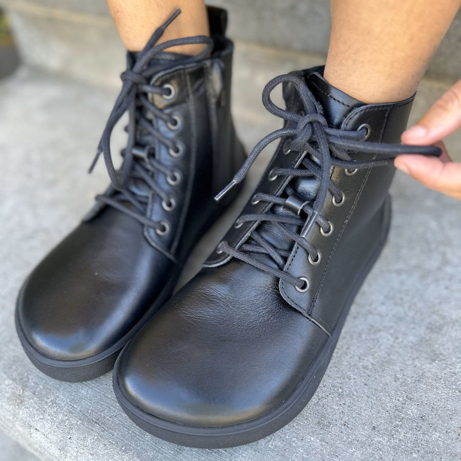 A photo of Be Lenka Atlas ankle zip up boots made from smooth leather and rubber soles. The boots are black in color with black laces, zippers, a pull tab in the back, and lined with felt. Both boots are shown diagonally from the front left on feet sitting on cement stairs. #color_black