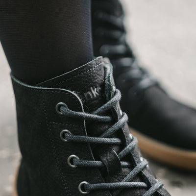 A photo of Be Lenka Atlas ankle zip up boots made from nubuck leather and rubber soles. The boots are black in color with black laces, zippers, a pull tab in the back, and lined with felt. Both boots are shown on a woman's feet with a view of her shins down and a close up of the laces of the right boot. The woman is wearing sheer black tights with the boots. #color_matte-black