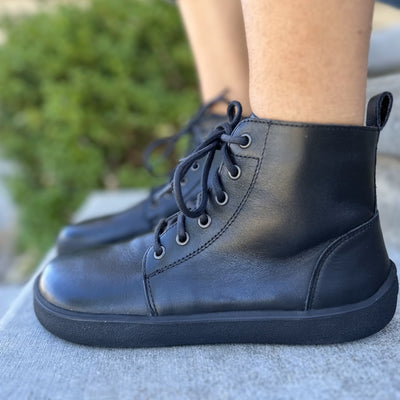 A photo of Be Lenka Atlas ankle zip up boots made from smooth leather and rubber soles. The boots are black in color with black laces, zippers, a pull tab in the back, and lined with felt. Both boots are shown from the left side on feet sitting on cement stairs with a green bush in the background. #color_black