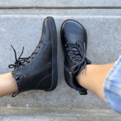 A photo of Be Lenka Atlas ankle zip up boots made from smooth leather and rubber soles. The boots are black in color with black laces, zippers, a pull tab in the back, and lined with felt. Both boots are shown from the top down on feet wearing light cropped jeans sitting on cement stairs. Left foot it leaning to the left to show the zipper. #color_black