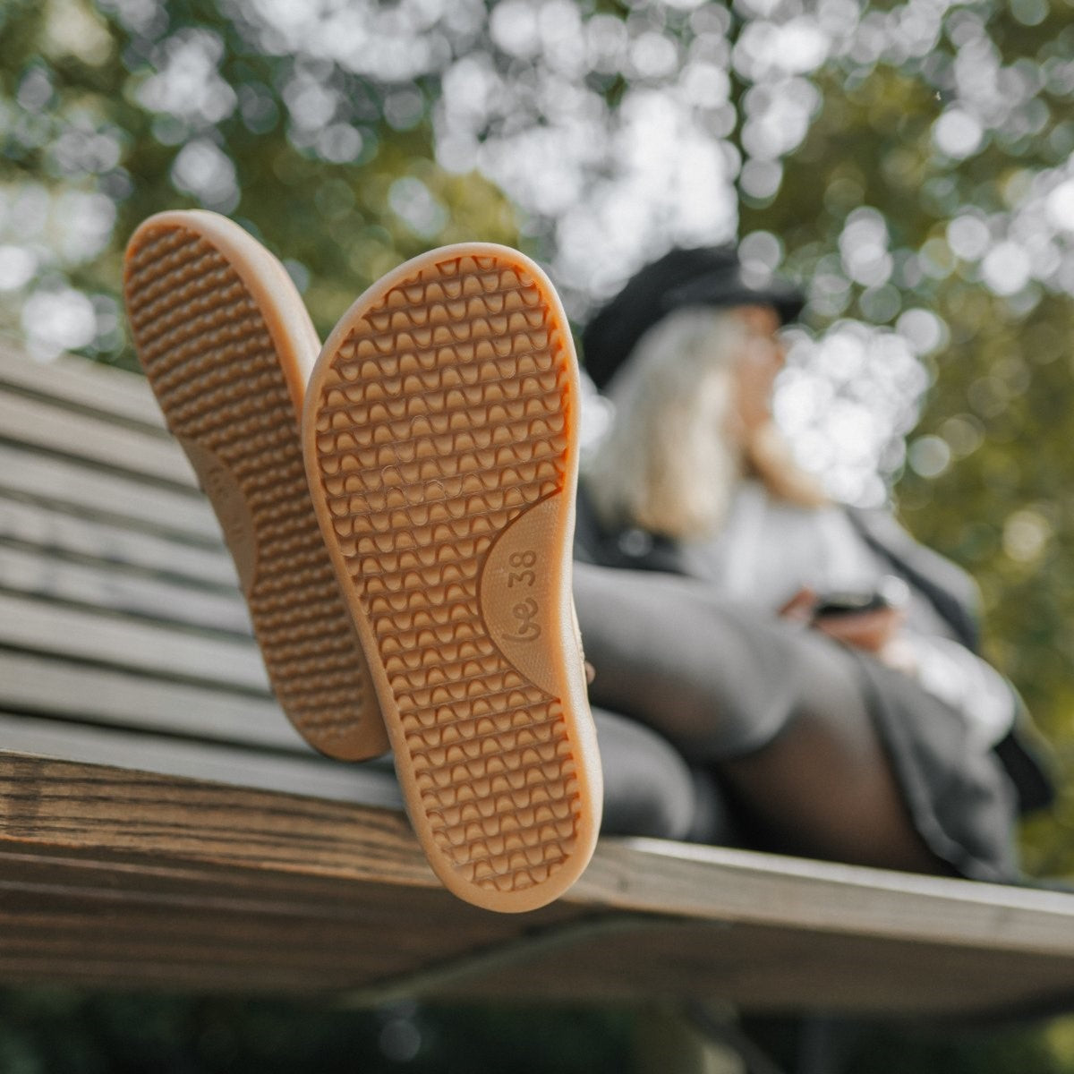 A photo of Be Lenka Atlas ankle zip up boots made from smooth leather and rubber soles. The boots are chocolate in color with black laces, zippers, a pull tab in the back, and lined with felt. The boots are shown from the soles on a woman's fee. The woman is sitting on a park bench with her feet up and is wearing a black outfit and a black hat. #color_chocolate