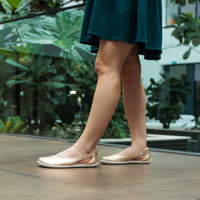 A photo of Be Lenka Bellissima flats with a leather upper and rubber soles. The flats are gold in color with a small stitching V detail in the front and a cut out design on the sides. Both shoes are shown from the left side on a woman wearing an above-the-knee teal dress with plants in the background. #color_gold