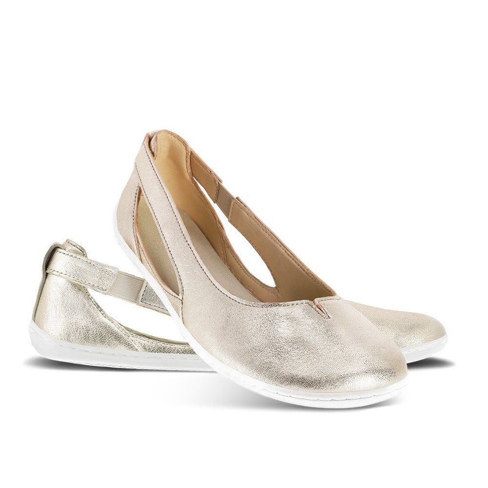 A photo of Be Lenka Bellissima flats with a leather upper and rubber soles. The flats are gold in color with a small stitching V detail in the front and a cut out design on the sides. Left shoe is shown from the right side with the right shoe propped up on the left shoe against a white background in this photo. #color_gold