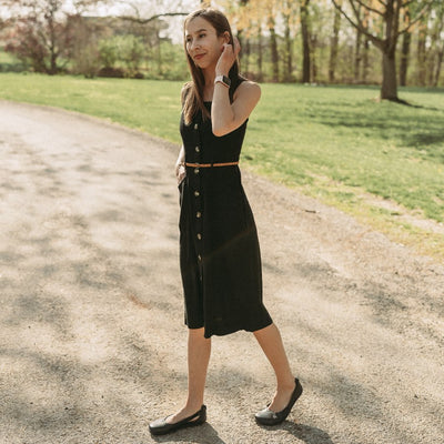 A photo of Be Lenka Bellissima flats with a leather upper and rubber soles. The flats are a black color with a small stitching V detail in the front and a cut out design on the sides. Shoes are shown from the left side on a woman with long brown hair standing on a country road with green grass and trees behind wearing a black thick strap knee length tank top dress with brown buttons down the front and a thin brown belt around the waist. #color_black