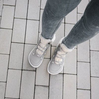 A photo of Belenka Bliss Boots made from nubuck leather and light grey rubber soles. The boots are grey and have fleece surrounding the whole tongue and around the ankle. The laces are wide across the boot top and have speed hooks at the by the top. Both boots are shown from the top down on a woman wearing grey skinny jeans. Woman is standing on a grey rectangle brick pathway. #color_cloud-grey