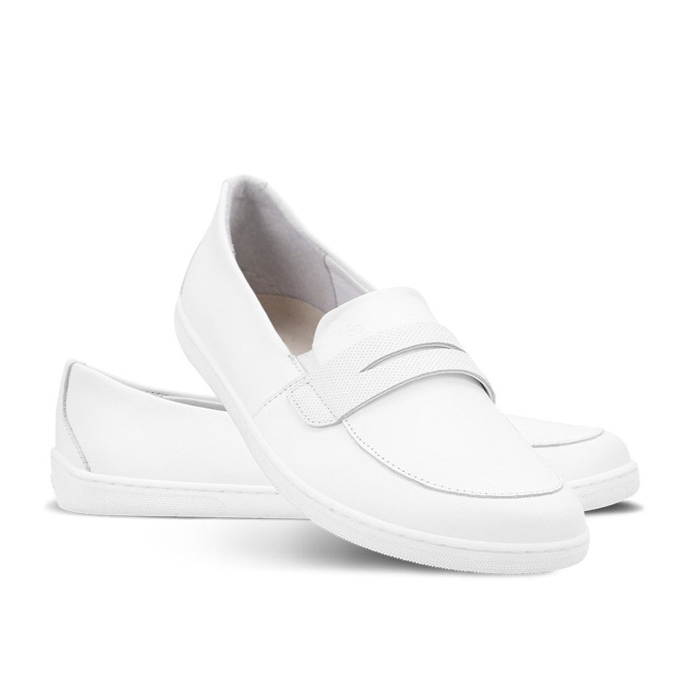 Photo 1 - A photo of Be Lenka Buena penny loafer in all white. The left loafer is shown from the side facing towards the right with the right shoe heel leaning against it against a white background. Photo 2 - Both shoes are shown from the front on feet against a brick housing complex background. #color_all-white