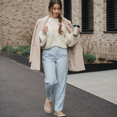 A photo of a penny loafer with a decorative vamp. A dark haired woman with a braid is shown wearing a white sweater, tan jacket resting on her shoulders, blue pants, and the loafers while she holds a coffee mug. There is a tan brick building behind her with greenery, and she walks on pavement. #color_latte-brown