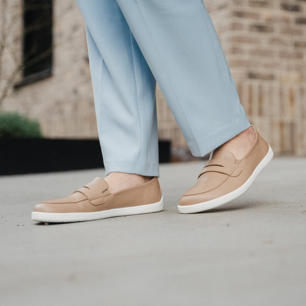 A photo of a penny loafer with a decorative vamp. A woman’s legs are shown from the knee down wearing blue pants and the loafers while she faces towards the left. She stands on concrete while a tan brick building is in the background. #color_latte-brown