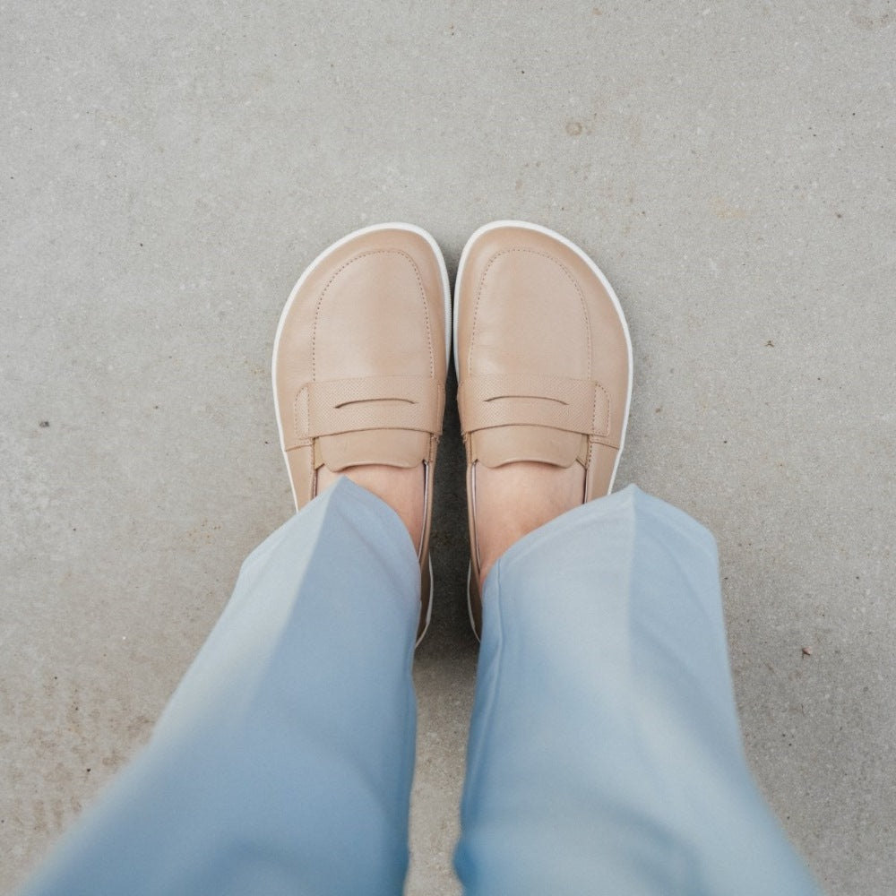 A photo of a penny loafer with a decorative vamp. A woman is shown standing with her feet facing forward while wearing blue pants and the loafers standing on concrete. #color_latte-brown
