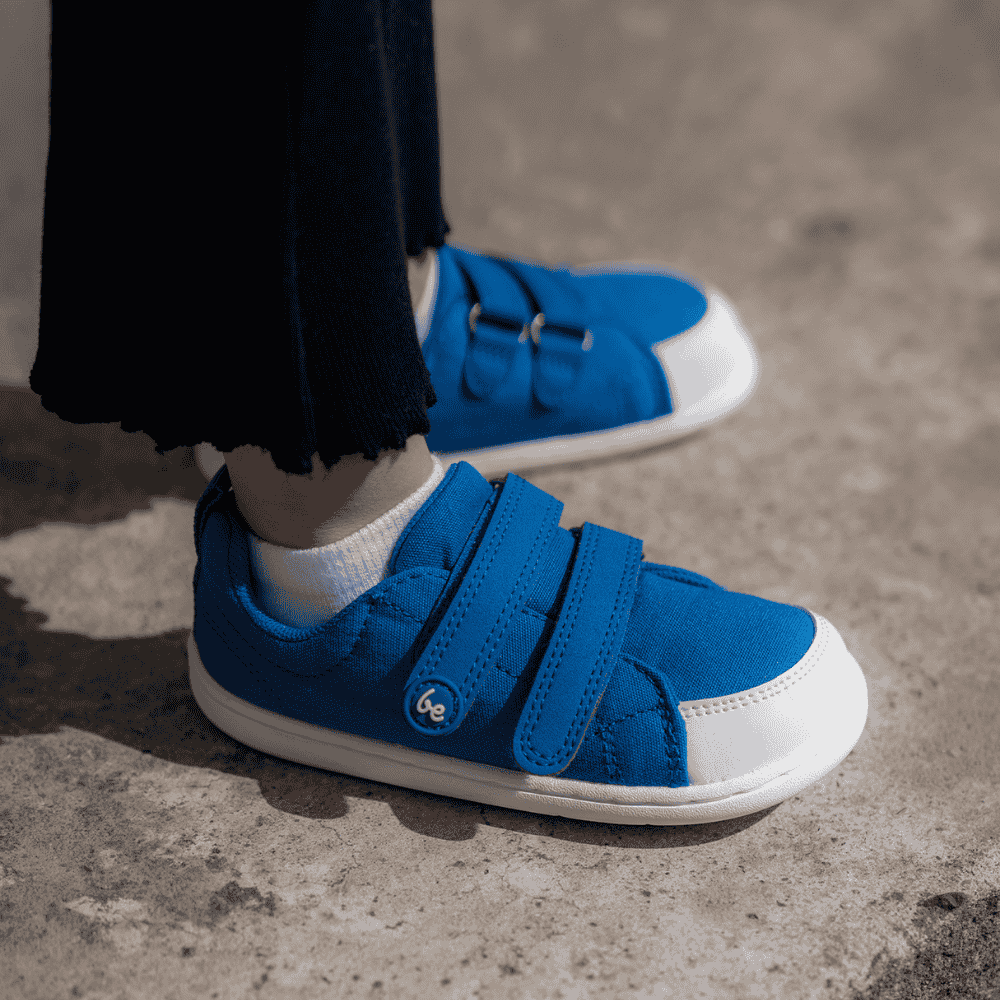 Blue canvas Be Lenka Canvi sneakers with  two velcro straps, white faux suede toe caps, white and teal rubber soles, and a pull tab at the heel. Both shoes are shown from the side facing right on a little girl wearing black pants standing on cement. #color_blue