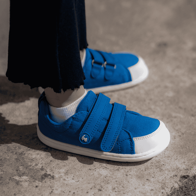 Blue canvas Be Lenka Canvi sneakers with  two velcro straps, white faux suede toe caps, white and teal rubber soles, and a pull tab at the heel. Both shoes are shown from the side facing right on a little girl wearing black pants standing on cement. #color_blue