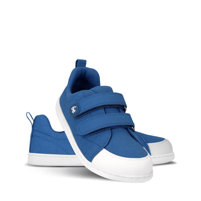 Blue canvas Be Lenka Canvi sneakers with  two velcro straps, white faux suede toe caps, white and teal rubber soles, and a pull tab at the heel. Left shoe is shown facing right with the right shoe heel propped up on it against a white background. #color_blue