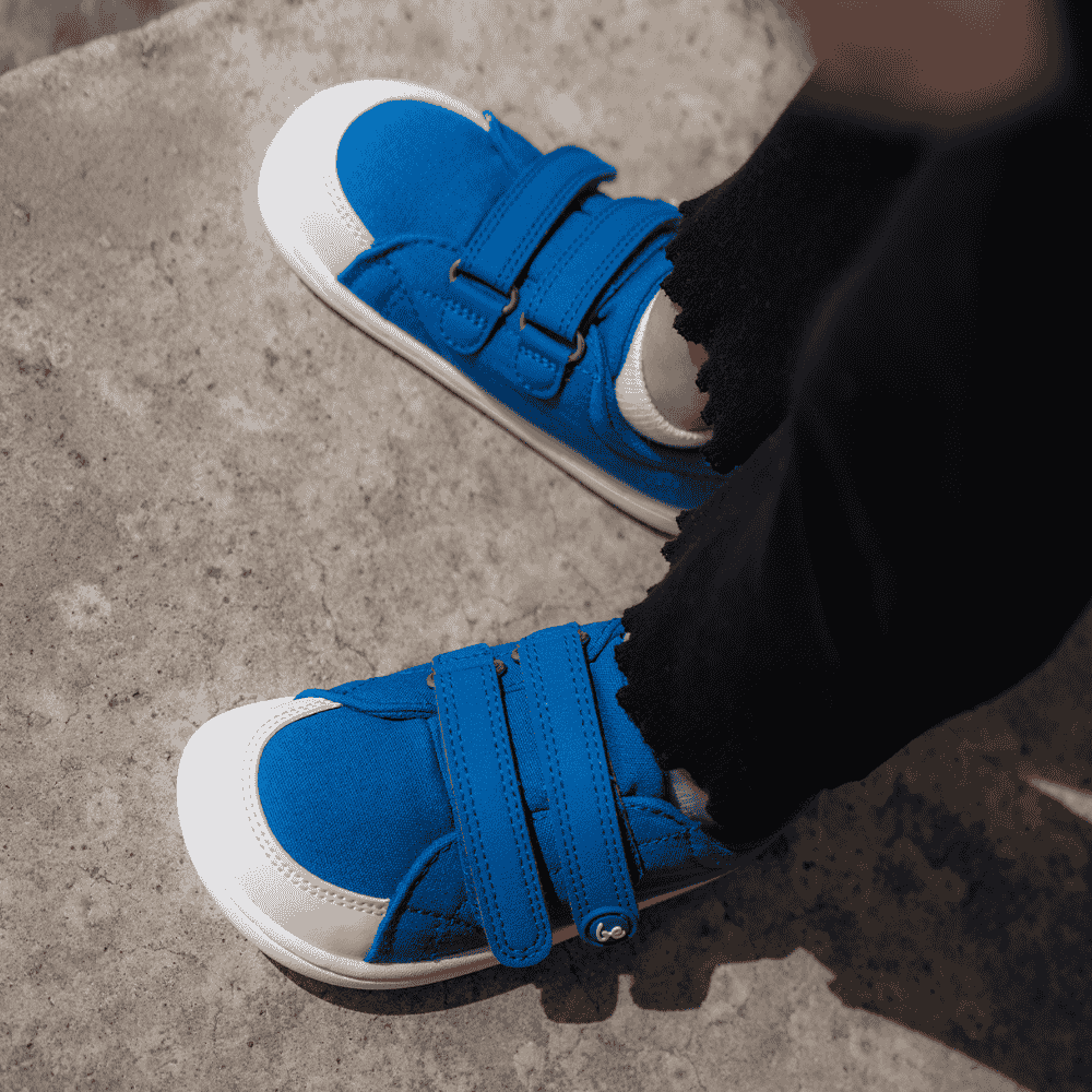 Blue canvas Be Lenka Canvi sneakers with  two velcro straps, white faux suede toe caps, white and teal rubber soles, and a pull tab at the heel. Both shoes are shown from above on a little girl facing left standing on cement. #color_blue