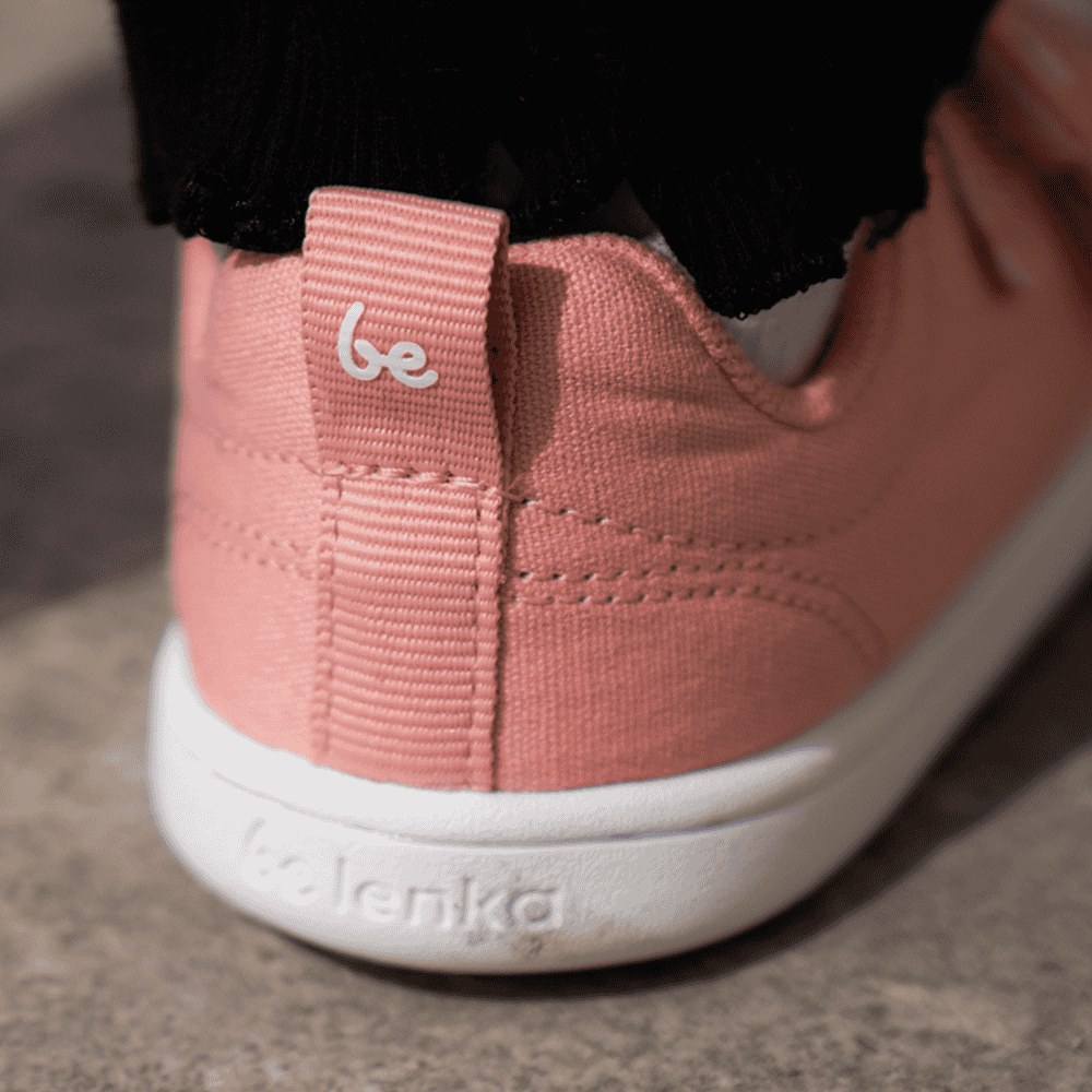 Light coral pink canvas Be Lenka Canvi sneakers with two velcro straps, white faux suede toe caps, white and coral pink rubber soles, and a pull tab at the heel. Right shoe heel is shown close up on a little girl standing on cement. #color_coral-pink