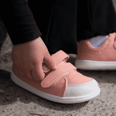 Light coral pink canvas Be Lenka Canvi sneakers with two velcro straps, white faux suede toe caps, white and coral pink rubber soles, and a pull tab at the heel. Both shoes are shown facing right on a little girl sitting on a cement stair closing a strap on the right shoe. #color_coral-pink