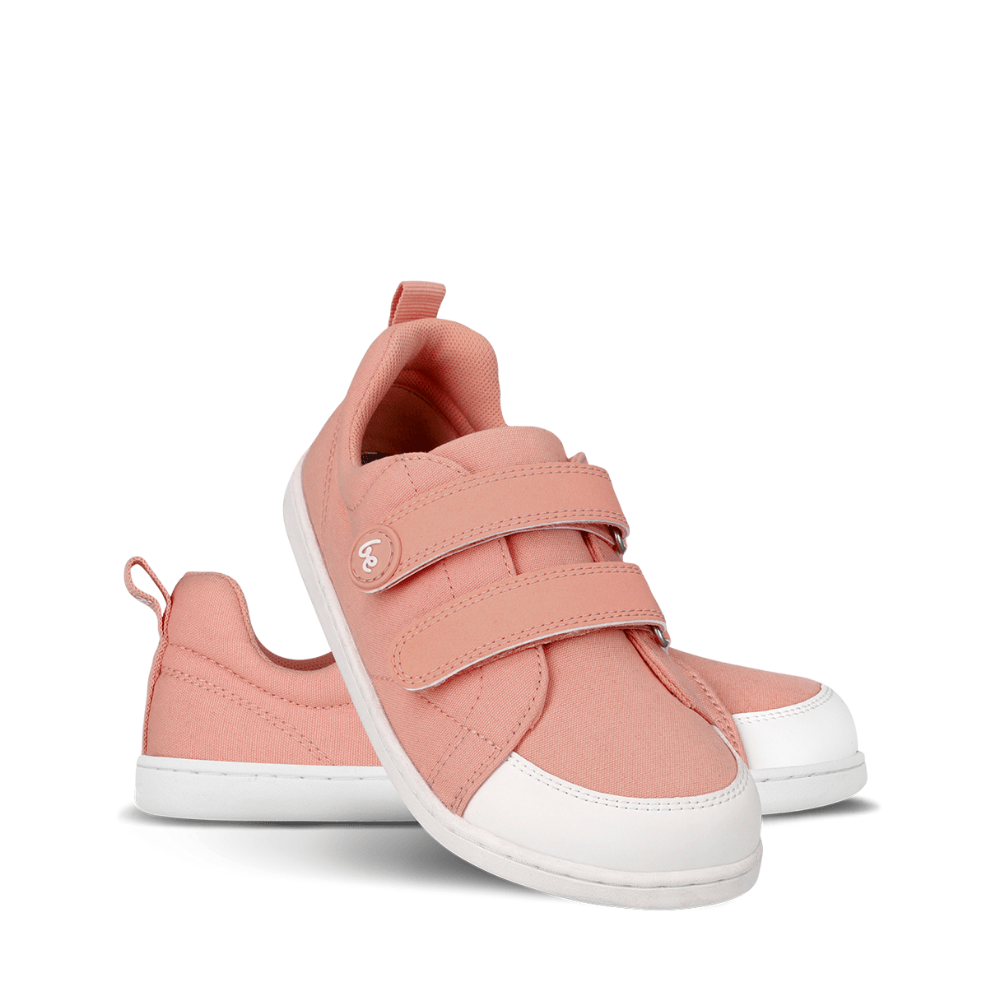 Light coral pink canvas Be Lenka Canvi sneakers with two velcro straps, white faux suede toe caps, white and coral pink rubber soles, and a pull tab at the heel. Left shoe is shown facing right with the right shoe heel propped up on it against a white background. #color_coral-pink