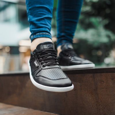 A photo of Belenka Champ sneakers made from leather and white rubber soles. The sneakers are black in color with perforation on the toe box and detail stitching. Both sneakers are shown from the front on a woman's feet with a view of her knees down. The woman is wearing blue skinny jeans and is standing on a brown step with her right foot in front of her left. #color_black-white