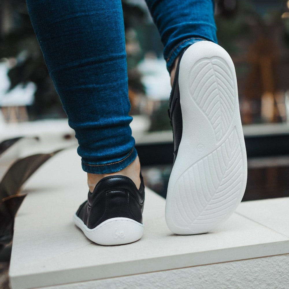 A photo of Belenka Champ sneakers made from leather and white rubber soles. The sneakers are black in color with perforation on the toe box and detail stitching. Both sneakers are shown from behind on a woman's feet with a view of her knees down. The woman is wearing blue skinny jeans and is walking on a white wall with her right foot up, showing the sole of the sneaker. #color_black-white