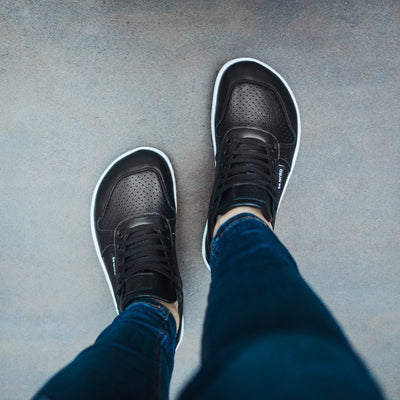 A photo of Belenka Champ sneakers made from leather and white rubber soles. The sneakers are black in color with perforation on the toe box and detail stitching. Both sneakers are shown from above on a woman's feet with a view of her knees down. The woman is wearing blue skinny jeans and standing on a grey cement floor. #color_black-white