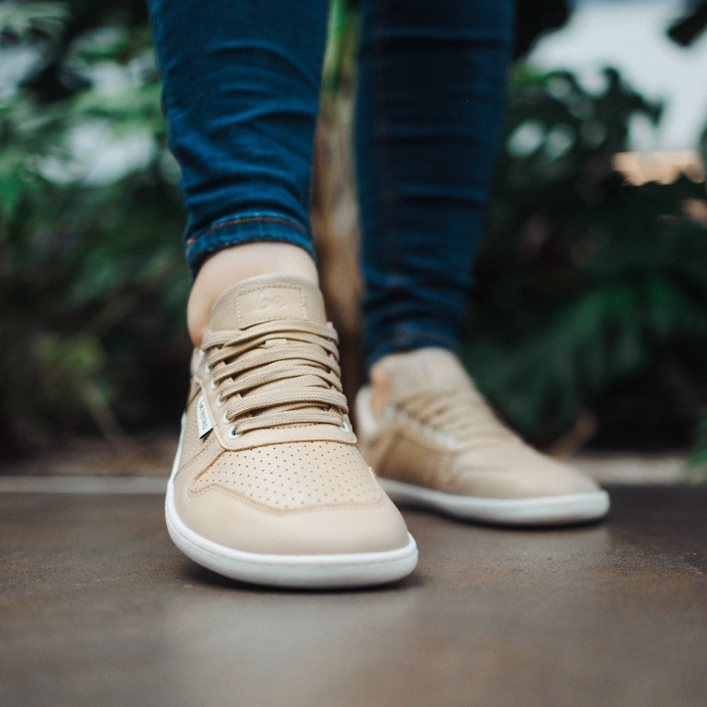 A photo of Belenka Champ sneakers made from leather and rubber soles. The sneakers are cappuccino in color with perforation on the toe box and detail stitching. Both sneakers are shown from the front on a woman's feet with a view of her knees down. The woman is wearing blue skinny jeans and is standing on a brown floor with greenery in the background. #color_cappuccino