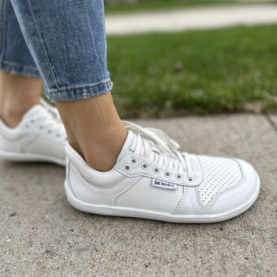 A photo of Be Lenka Champ sneakers made from leather and white rubber soles. The sneakers are white in color with perforation on the toe box and detail stitching. Both shoes are shown on a woman's feet from the right side with a view of her shins down. The woman is wearing cropped blue jeans and is standing on a paved sidewalk with her right foot in front of her left. #color_white