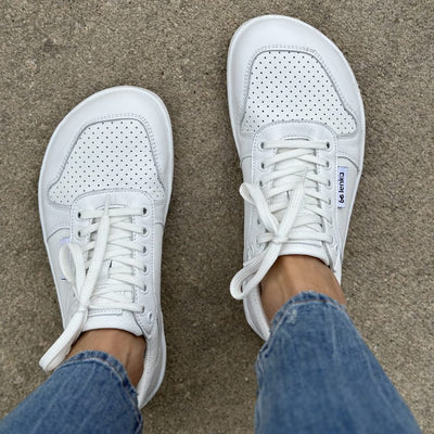 A photo of Be Lenka Champ sneakers made from leather and white rubber soles. The sneakers are white in color with perforation on the toe box and detail stitching. Both shoes are shown together from above on a woman's feet with a view of her shins down. The woman is wearing copped blue jeans and is standing on pavement.. #color_white