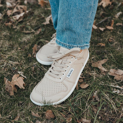 A photo of Be Lenka Champ sneakers made from leather and white rubber soles. The sneakers are cappuccino in color with perforation on the toe box and detail stitching. Both shoes are shown facing diagonally left on a woman wearing loose jeans standing on leaf covered grass. #color_cappuccino