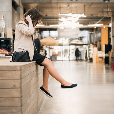 A photo of Black Be Lenka Delight knitted flats with white soles. Both shoes are shown facing right on a woman sitting on a wooden stage kicking her right leg out. She is wearing a black dress and a tan blazer with an urban indoor setting in the background. #color_black