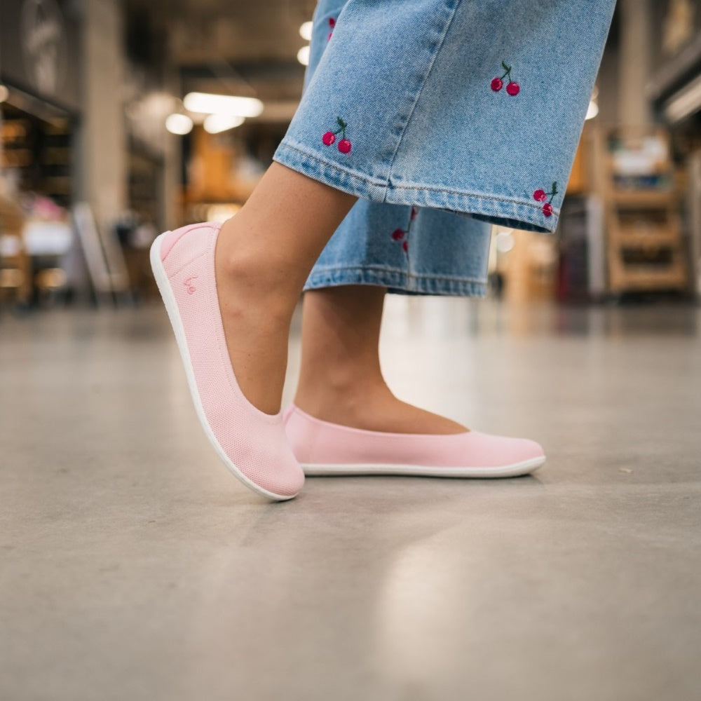 A photo of Light Pink Be Lenka Delight knitted flats with white soles. A woman wearing cropped, light-wash, wide-leg jeans with cherries embroidered on them is wearing these. She is standing on polished cement facing right with her right foot resting on the toe behind the left with an urban setting in the background. #color_light-pink