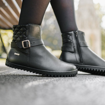 A photo of Be Lenka Diva boots in all black. Boots are made from smooth black leather. A quilted leather design is present in the back, a silver buckle on the side, a strap around the ankle, and a zipper. Both boots are shown from the right on a woman's feet. The woman is wearing black stockings and is walking on a paved road. #color_all-black