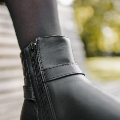 A photo of Be Lenka Diva boots in all black. Boots are made from smooth black leather. A quilted leather design is present in the back, a silver buckle on the side, a strap around the ankle, and a zipper. The left boot is shown from the right on a woman's foot, with a close up of the zipper on the inner ankle. The woman is wearing black stockings and is standing on a paved road. #color_all-black