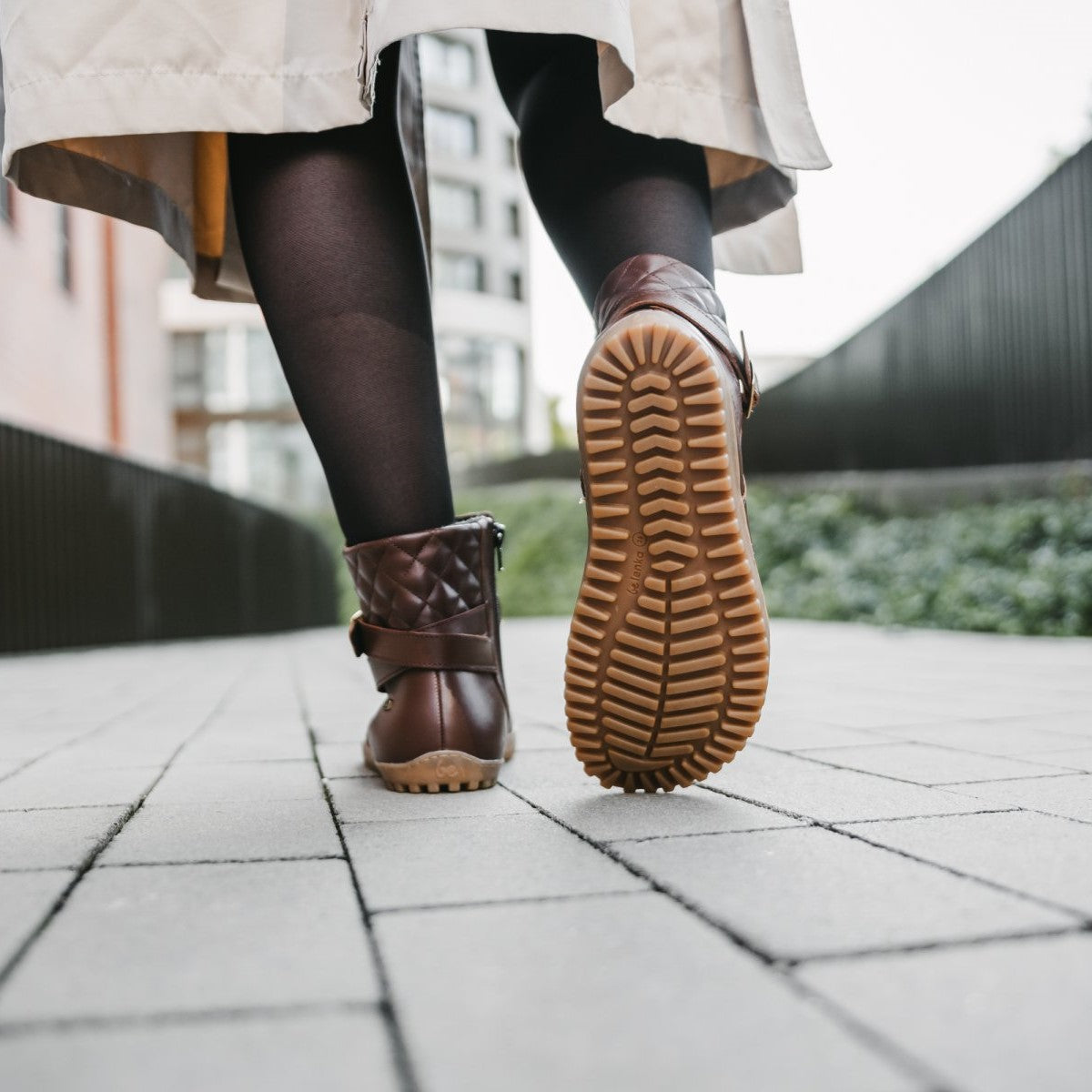 A photo of Be Lenka Diva boots in dark brown with tan soles. Boots are made from dark brown leather. A quilted leather design is present in the back, a gold buckle on the side, a strap around the ankle, and a zipper. The left shoe is shown from the back and the right shoe from the bottom on a woman's feet. The woman is wearing black stockings and a khaki trench coat and is walking on a paved sidewalk. #color_dark-brown