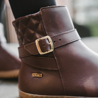 A photo of Be Lenka Diva boots in dark brown with tan soles. Boots are made from dark brown leather. A quilted leather design is present in the back, a gold buckle on the side, a strap around the ankle, and a zipper. The right boot is shown from the right on a woman's foot with a close up view of the quilting detail and the buckle. The woman is wearing black stockings and is standing on a paved sidewalk. #color_dark-brown