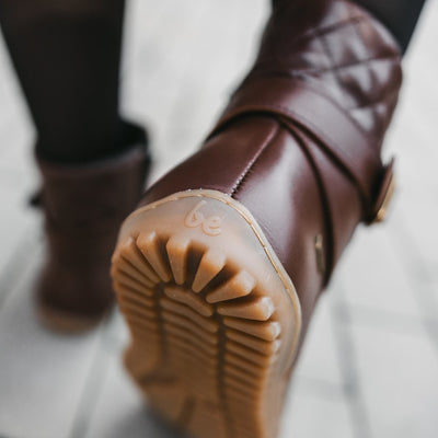 A photo of Be Lenka Diva boots in dark brown with tan soles. Boots are made from dark brown leather. A quilted leather design is present in the back, a gold buckle on the side, a strap around the ankle, and a zipper. Both shoes are shown from behind  on a woman's feet with a close up of the right heel. The woman is wearing black stockings and is standing on a paved sidewalk. #color_dark-brown