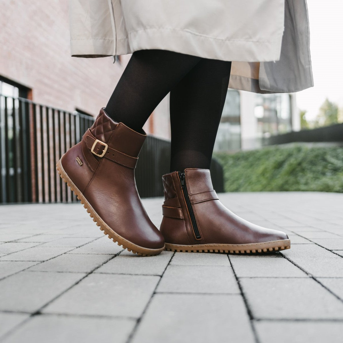 A photo of Be Lenka Diva boots in dark brown with tan soles. Boots are made from dark brown leather. A quilted leather design is present in the back, a gold buckle on the side, a strap around the ankle, and a zipper. Both shoes are shown from the right on a woman's feet. The woman is wearing black stockings and a khaki trench coat and is walking on a paved sidewalk. #color_dark-brown