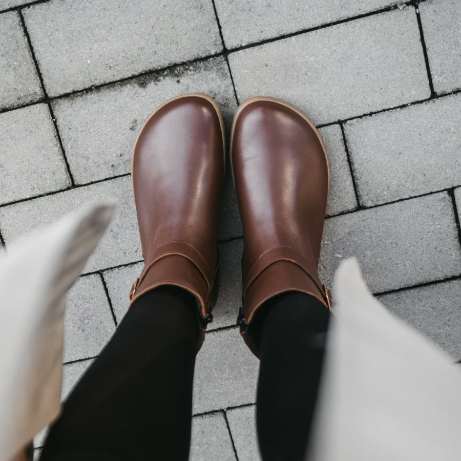 A photo of Be Lenka Diva boots in dark brown with tan soles. Boots are made from dark brown leather. A quilted leather design is present in the back, a gold buckle on the side, a strap around the ankle, and a zipper. Both shoes are shown from above on a woman's feet. The woman is wearing black stockings and a khaki trench coat and is standing on a paved sidewalk. #color_dark-brown