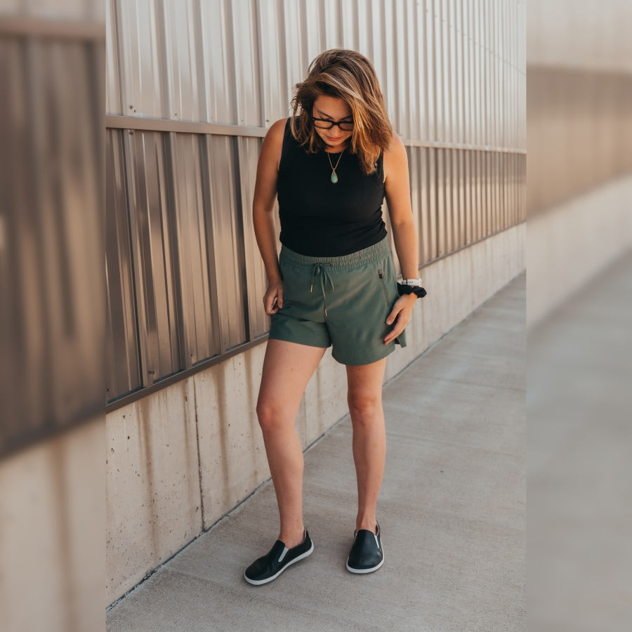 A photo of Belenka Eazy slip-on sneakers made from leather and rubber soles. The sneakers are a black color with white elastic on the sides and soles. A brown haired woman wearing glasses is shown standing on concrete in front of a metal and concrete building looking down with one leg out to the side and bent slightly. She is wearing a black tank top, green shorts, green pendant necklace, and the Eazy sneakers. #color_black