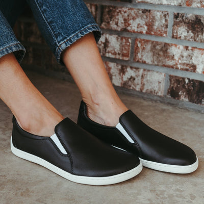 A photo of a black leather slip-on sneaker with white elastic on the sides, and a white sole. Both shoes are shown here facing diagonally right worn on a woman wearing cropped medium-wash blue jeans sitting on cement connected to a distressed brick wall. #color_black-white-leather