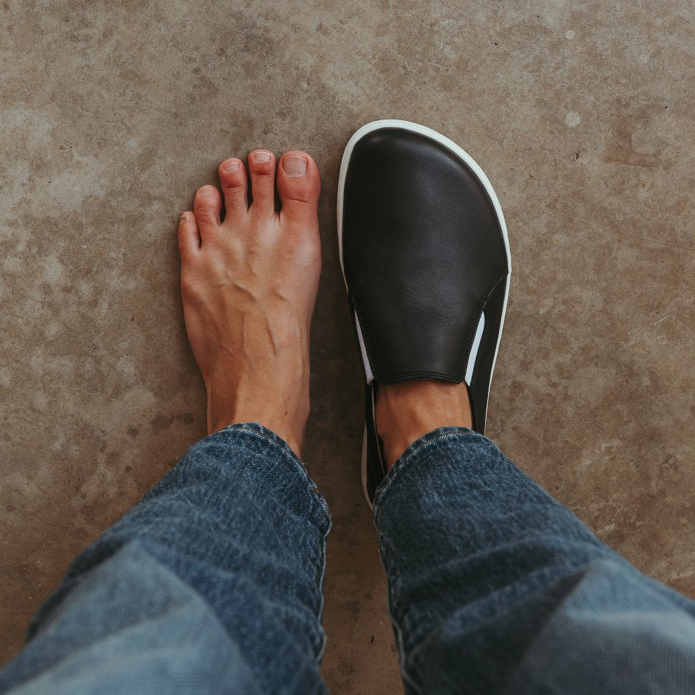 A photo of a black leather slip-on sneaker with white elastic on the sides, and a white sole. Right show is shown here from above worn on a woman wearing cropped medium-wash blue jeans standing on cement. The left foot is bare. #color_black-white-leather