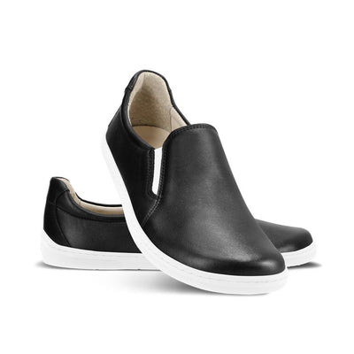 A photo of a black leather slip-on sneaker with white elastic on the sides, and a white sole. Both sneakers are shown the right sneaker is shown from the front while resting its heel on the left sneaker behind it against a white background. #color_black-white-leather