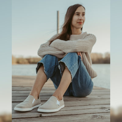 A photo of a sand canvas slip-on sneaker with white elastic on the sides, and a white sole. Both sneakers are shown here from the front facing diagonally left. They are worn on a tan woman with long, straight, brown hair wearing a sand knit sweater and light-wash, cropped, wide-legged jeans. She is sitting on a wooden dock with water and a tree line in the background. #color_sand-canvas