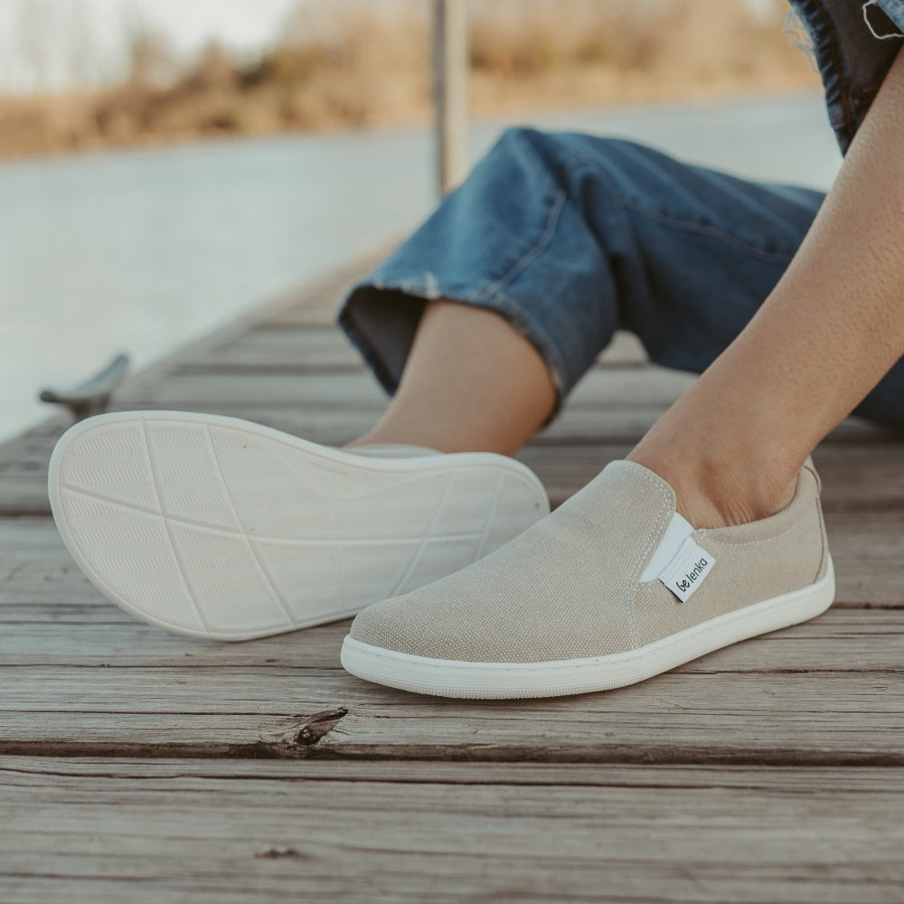 A photo of a sand canvas slip-on sneaker with white elastic on the sides, and a white sole. Both sneakers are shown here from the side facing diagonally left with the right shoe sole facing the camera. They are worn on a woman wearing light-wash, cropped, wide-legged jeans. She is sitting on a wooden dock with water and a tree line in the background. #color_sand-canvas