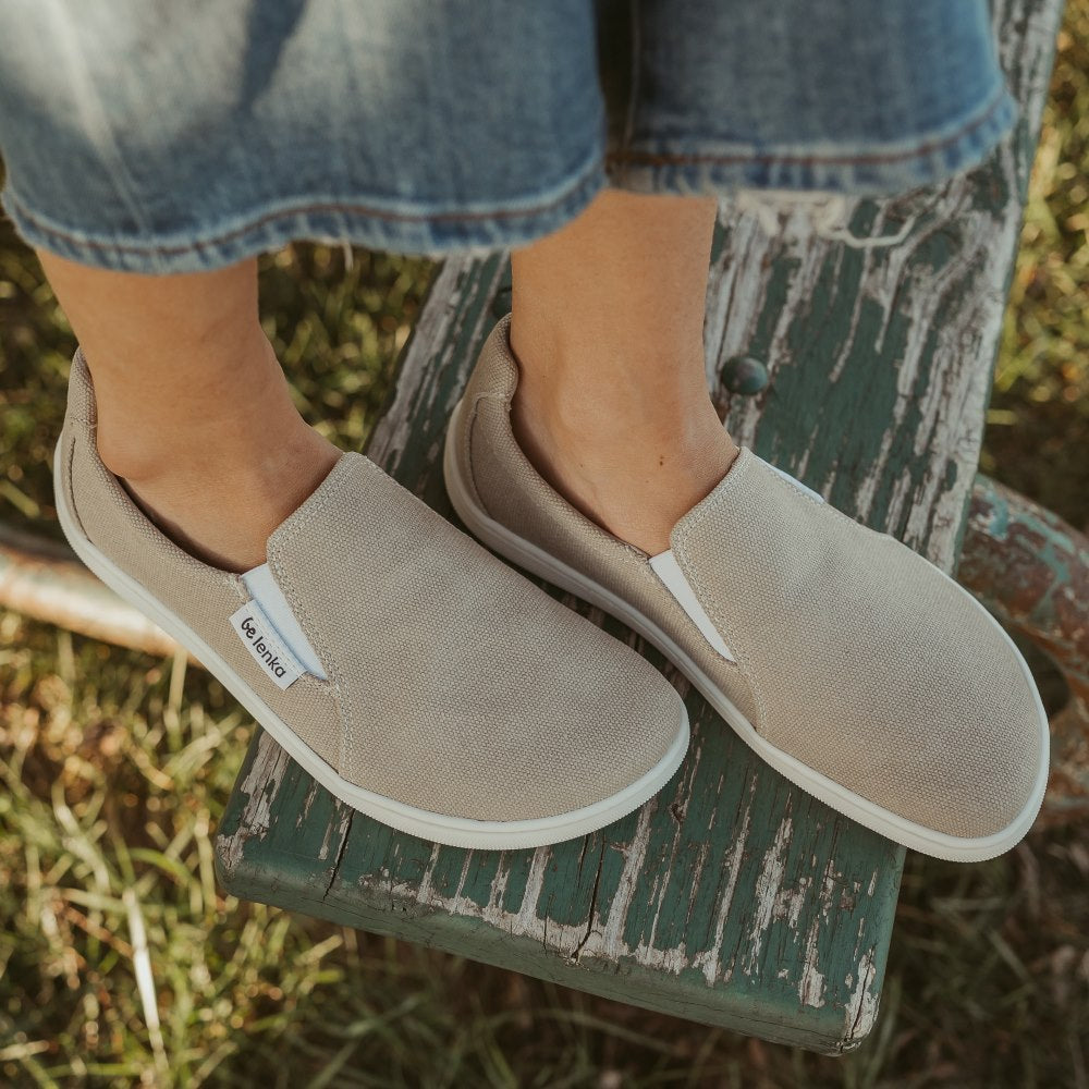 A photo of a sand canvas slip-on sneaker with white elastic on the sides, and a white sole. Both sneakers are shown here from above facing diagonally right. They are worn on a woman wearing cropped light-wash wide-legged jeans sitting on an old metal and wooden bench resting on grass. #color_sand-canvas