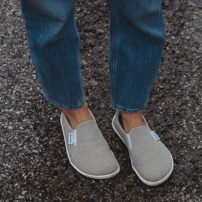 A photo of Belenka Eazy slip-on sneakers made from canvas and rubber soles. The sneakers are a sand color with white elastic on the sides and soles. A woman is shown from knee down facing forward standing on a gravel road wearing jeans and sand eazy sneakers. #color_sand