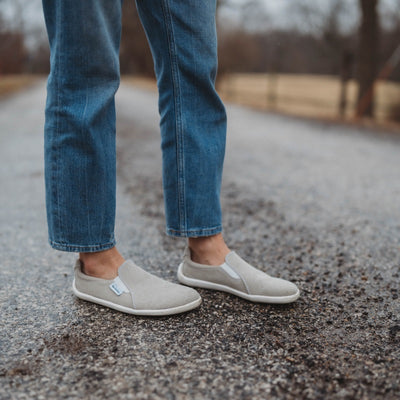 A photo of Belenka Eazy slip-on sneakers made from canvas and rubber soles. The sneakers are a sand color with white elastic on the sides and soles. A woman is shown mid thigh down standing facing right on a gravel road with blurry trees in the distance, she is wearing jeans and the the sand eazy sneakers. #color_sand